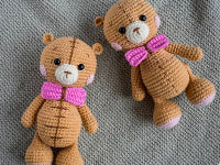 Crochet teddy bears with pink bows lying on fabric background, available at all-in-1store.com.