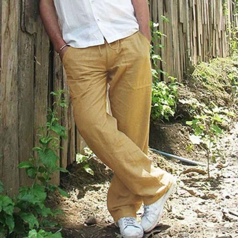 Man wearing khaki cotton linen trousers, casual style, leaning against a wooden fence in a natural outdoor setting.